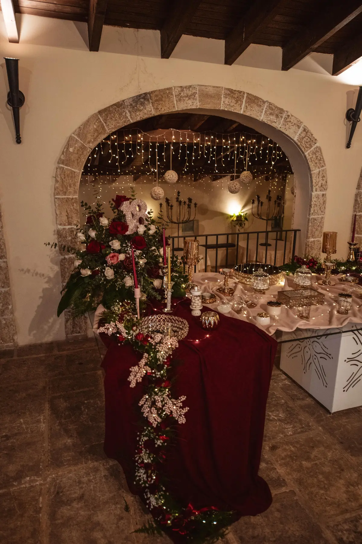 Il tavolo della torta col drappo bordeaux, i fiori e la cascata di lucine sotto l'arco